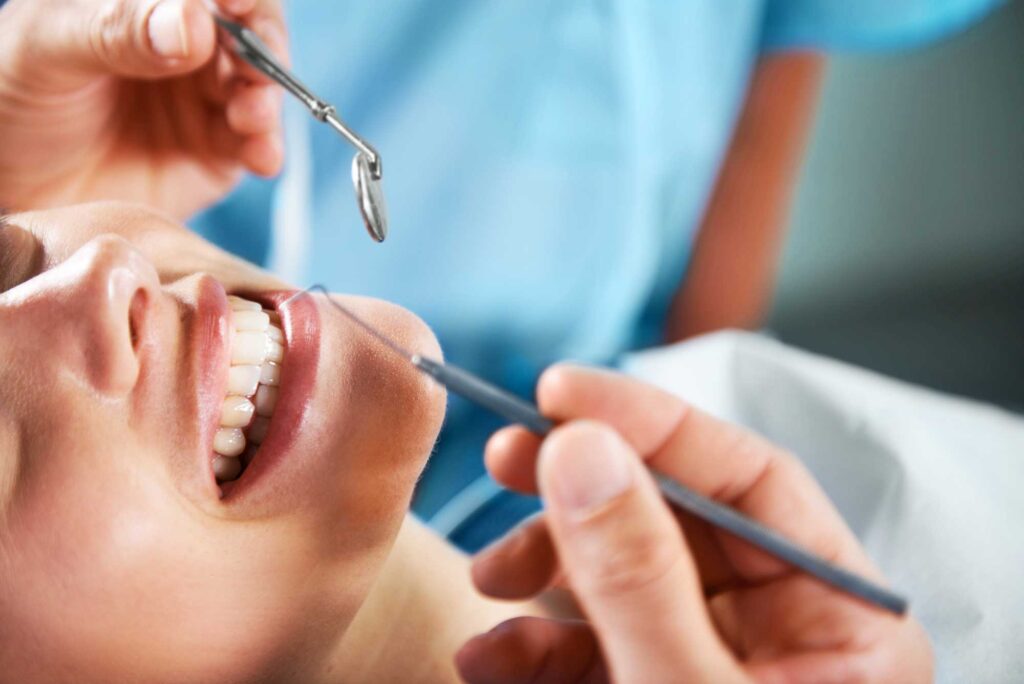 Close-up of a patient smiling during a dental examination, with a dentist holding a dental mirror and an explorer tool while examining the patient's teeth. The patient appears relaxed and comfortable.