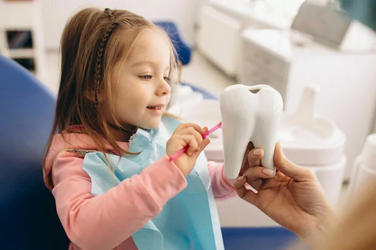 A young girl in a dental clinic holding a pink toothbrush and brushing a large tooth model held by a dental professional. The girl is wearing a blue dental bib and smiling, demonstrating proper tooth brushing techniques. The background shows typical dental office equipment.