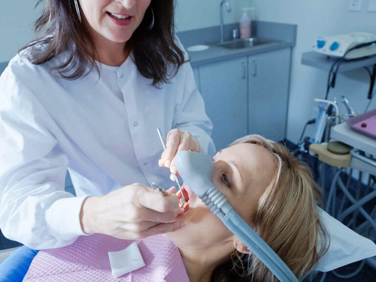 A dental professional treating a patient in a dental chair. The patient is wearing a nasal mask for nitrous oxide sedation, commonly known as laughing gas, and a pink dental bib. The dentist is holding dental tools, performing a procedure in the patient's mouth. The background shows typical dental office equipment and a clean, clinical setting.
