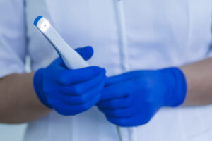 A dental professional wearing blue gloves holding an intraoral camera, which is used for taking detailed images of a patient's mouth. The camera is lit, indicating it is ready for use. The background shows the dental professional wearing a white coat.