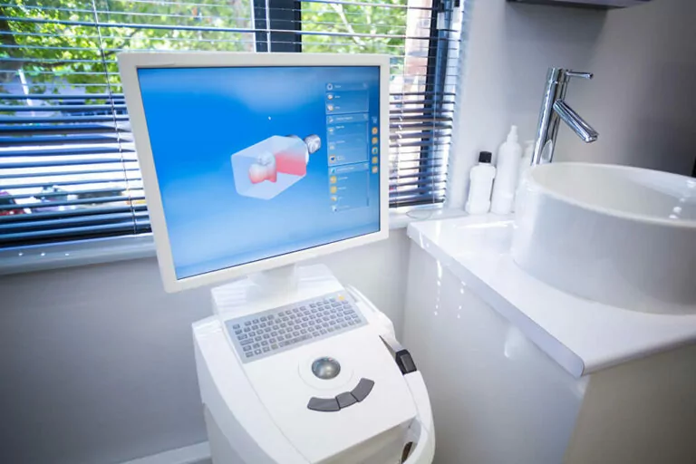 A computer monitor displaying dental imaging software with a 3D model of a tooth. The equipment is set up in a modern dental office, next to a sink and various dental care products. The window blinds in the background let in natural light, creating a bright and clean environment.